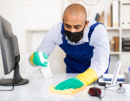 Man professional cleaner in medical mask cleaning desk with computer in office